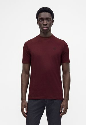 Ung mand iført en ensfarvet mørkerød kortærmet t-shirt og mørke bukser, stående mod en lysegrå baggrund.