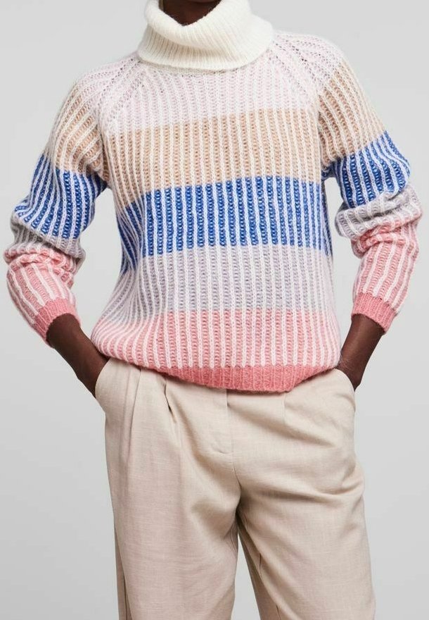Stickad tröja med hög krage, prydd med horisontella ränder i rosa, blått och vitt samt ribbat mönster. Beiga veckade byxor.