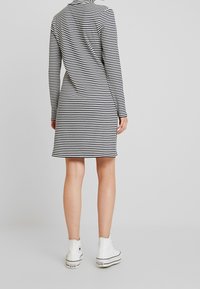 Czarna i biała sukienka w paski z długim rękawem, dopasowana, do kolan, wykonana z teksturowanej tkaniny. Uzupełniona białymi sportowymi butami za kostkę.