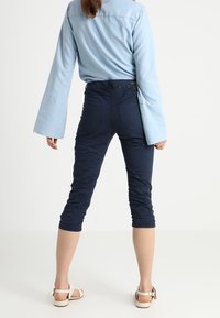 Kvinna i ljusblå topp med vida ärmar, marinblå cropped byxor och vita sandaler som står mot en enkel vit bakgrund.