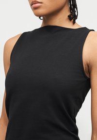 Gros plan sur une personne portant un haut noir sans manches à col montant, montrant le haut du torse et des mèches de cheveux tressés.