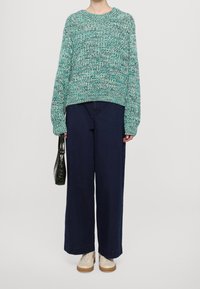 Türkisfarbener, strukturierter Strickpullover mit langen Ärmeln, kombiniert mit weiten, dunkelblauen Hosen und beigen Sneakers, während eine schwarze Handtasche gehalten wird.