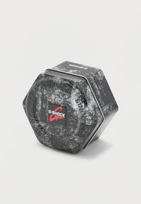 Εξάγωνο μεταλλικό κουτί με φθαρμένη μαύρη επιφάνεια, με λογότυπο G-SHOCK σε κόκκινο και λευκό. Μεταλλικό κλείσιμο στην πάνω άκρη.