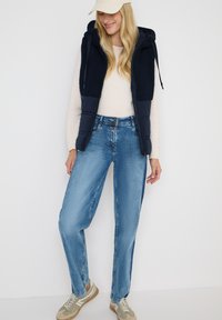 Chaleco acolchado azul marino con capucha de forro polar, suéter crema, jeans sueltos de color azul claro y zapatillas beige con acentos en azul claro.