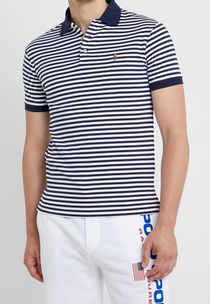 Homme portant un polo rayé bleu marine et blanc avec un short blanc présentant un petit drapeau américain et un texte bleu et rouge sur le côté.