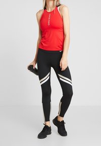 Débardeur sportif rouge sans manches avec le logo "Nike Pro", leggings noirs avec des rayures diagonales blanches et dorées, et chaussures de sport noires.
