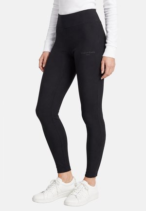 Mujer que lleva puestos leggings ajustados negros de cintura alta y zapatillas blancas, de pie con las manos relajadas a los lados sobre un fondo liso.