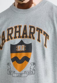 Grau Baumwoll-T-Shirt mit "CARHARTT" in Dunkelorange, einem Schildgrafik mit "WP" und "THE LAZY DUCK" sowie dekorativen Scroll-Akzenten.