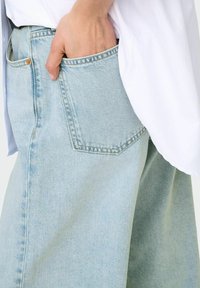 Jean en denim bleu clair avec une coupe décontractée, doté d'une poche arrière, de légères usures et d'une taille haute. Détail de tissu texturé visible.