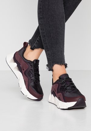 Person wearing black and dark maroon chunky sneakers with white soles and frayed black jeans, standing on a smooth gray floor.