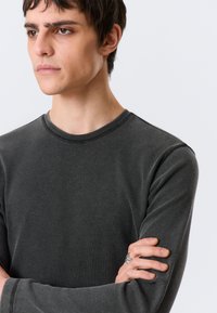 Σκούρο γκρι μακρυμάνικο πουκάμισο με ανάγλυφη υφή, στρογγυλή λαιμόκοψη και εμφανείς ραφές. Το μοντέλο ποζάρει με σταυρωμένα τα χέρια, φοράει δαχτυλίδι.