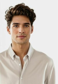 Ungdommelig mann med krøllete mørkt hår og lys beige skjorte med knapper, som ser direkte inn i kameraet med et nøytralt uttrykk.