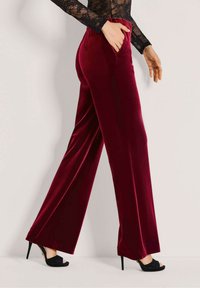 Vrouw in bordeauxrode fluwelen broek met hoge taille en wijde pijpen, zwart open teenhakken, hand in zak, tegen een effen lichte achtergrond.
