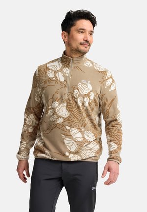 Mann trägt einen beige-weiß gemusterten Fleece-Pullover mit Reißverschlusskragen und dunkle Hose, schaut leicht zur Seite.