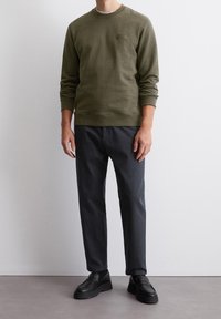 Oliwkowa bluza dresowa z okrągłym dekoltem i ściągaczami na rękawach, zestawiona z ciemnoszarymi spodniami o zwężanym kroju i czarnymi, casualowymi butami. Minimalistyczny design.