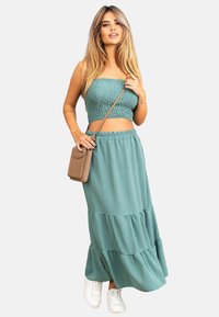 Teal to-delt sett bestående av en smocked crop top og et lagdelt maxi skjørt. Materialet er mykt med en jevn tekstur; kombinert med hvite joggesko.