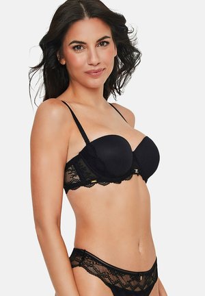 Femme portant un soutien-gorge en dentelle noire et une culotte assortie en dentelle avec des détails transparents, posant sur un fond clair uni.
