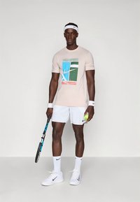 Lys rosa t-skjorte med korte ermer og grafisk trykk, kombinert med hvite shorts. Figuren holder en tennisracket og ball, og har på seg hvite joggesko.