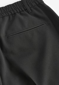 Pantalones negros de tejido suave con una cintura elástica, que cuentan con un único bolsillo trasero con cremallera y un diseño limpio y minimalista.