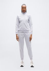 Αθλητική φόρμα σε ανοιχτό γκρι χρώμα, αποτελούμενη από μπουφάν με φερμουάρ και παντελόνι με στενή γραμμή, κατασκευασμένη από λείο ύφασμα με διακριτική λάμψη. Διακοσμημένη με λευκό λογότυπο.