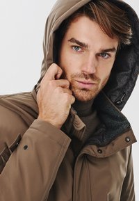 Brązowa, ocieplana kurtka z teksturowanym szarym podszyciem i regulowanym kapturem. Zawiera zamek błyskawiczny i guziki na rękawach.