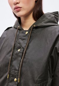 Tume roheline vahatatud jakk kapuutsiga, millel on kuldsed trukid, pruun reguleeritav nöör ja tekstureeritud kangas robustse välimuse jaoks.