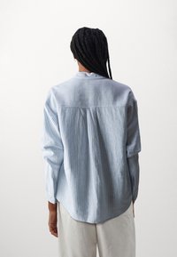 Ljusblå randig skjorta, långa ärmar, detaljer med veck bak, rundad nederkant, mjuk textur, avslappnad stil. Vita byxor delvis synliga.