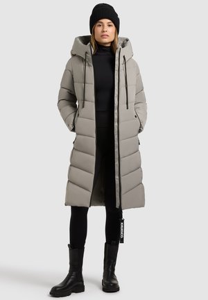 Langer grauer Daunenmantel mit Kapuze, ausgestattet mit einem gesteppten Design, Reißverschluss und seitlichen Taschen. Kombiniert mit schwarzen Leggings und Ankle Boots.