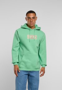 Mężczyzna z piegami noszący miętowozieloną bluzę z kapturem z naszywką w kształcie dolara i niebieskie dżinsy, stojący na szarym tle.