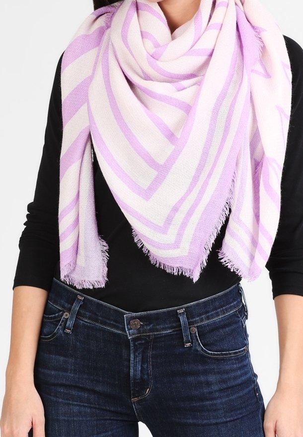 Personne portant une grande écharpe triangulaire rayée rose et blanche avec des bords à franges, par-dessus un haut noir et un jean bleu.