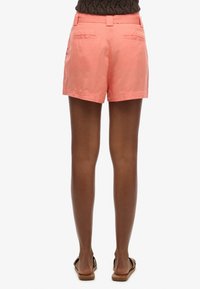 Coral pink shorts with a smooth texture, two back pockets, and a fitted waist. Paired with brown sandals, view from the back.