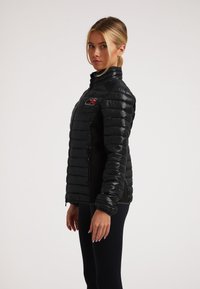 Giacca a vento nera con finitura lucida, chiusura frontale con zip, polsini a coste e una toppa con logo sul lato sinistro del petto. Presenta una silhouette aderente.
