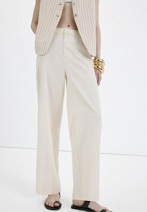 Persona con pantalones beige de pierna ancha, un chaleco sin mangas a rayas, pulseras doradas y sandalias negras con dedos abiertos, de pie frente a un fondo blanco.