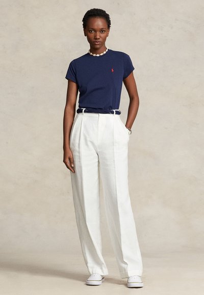 Γυναίκα που φοράει μπλε ναυτικό μπλουζάκι, λευκό παντελόνι με φαρδύ πόδι και ψηλή μέση, λευκά αθλητικά παπούτσια και κολιέ με κοχύλια, στέκεται με το ένα χέρι στην τσέπη.