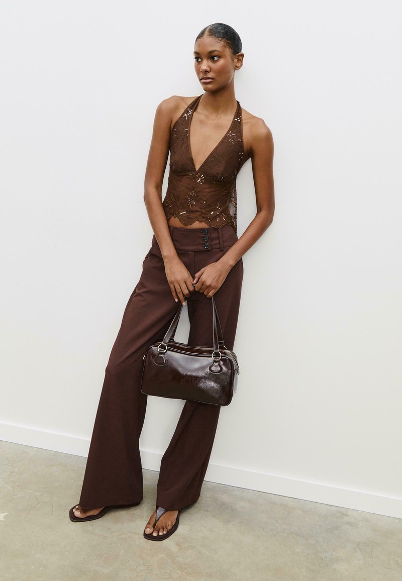 Hnědý krajkový halter top s květinovou výšivkou, spárovaný s hnědými kalhotami s vysokým pasem a lesklou hnědou kabelkou. Sandály jsou také hnědé.