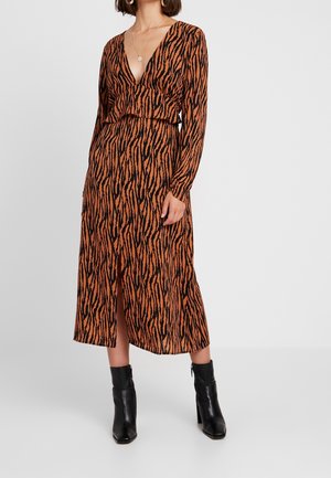 Vrouw in een lange mouwen, bruin en zwart dierenprint jurk met een diepe V-hals en zwarte high-heeled enkelboots.
