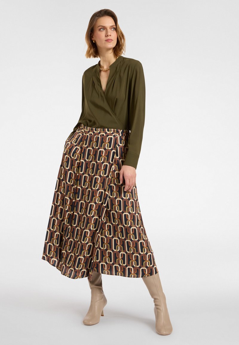 Blusa avvolgente verde oliva abbinata a una gonna maxi marrone con motivo a forme intrecciate in beige e arancione. Modella con stivaletti beige alla caviglia.