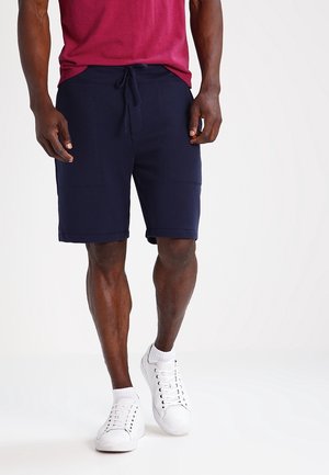 Homme portant un short bleu marine à cordon, des baskets blanches, des chaussettes blanches et une chemise rouge à manches courtes marchant devant un fond uni.