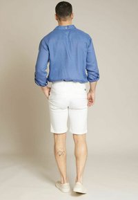 Chemise en lin bleu clair avec les manches retroussées, shorts blancs et chaussures de couleur claire. Tatouage notable visible sur la jambe droite.
