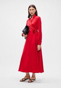 Czerwona sukienka z długimi rękawami, ozdobną różą w talii i zwiewnym dołem. Noszona z czarną kopertówką i płaskimi sandałami.