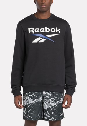 Sudadera negra de Reebok con un gran logo en blanco y azul. Presenta un cuello redondo y puños acanalados. Combinada con pantalones cortos de patrón mármol en blanco y negro.