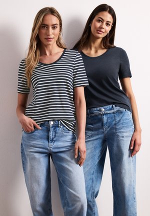 Dwie kobiety stoją przy białej ścianie, ubrane w krótkie topy — jedna w paski granatowo-białe, druga jednokolorowa granatowa — i jasnoniebieskie dżinsy.