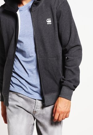 Mężczyzna ubrany w ciemnoszarą bluzę z kapturem na zamek z białym logo na piersi, pod którą ma jasnoniebieski T-shirt oraz jasnoszare dżinsy.
