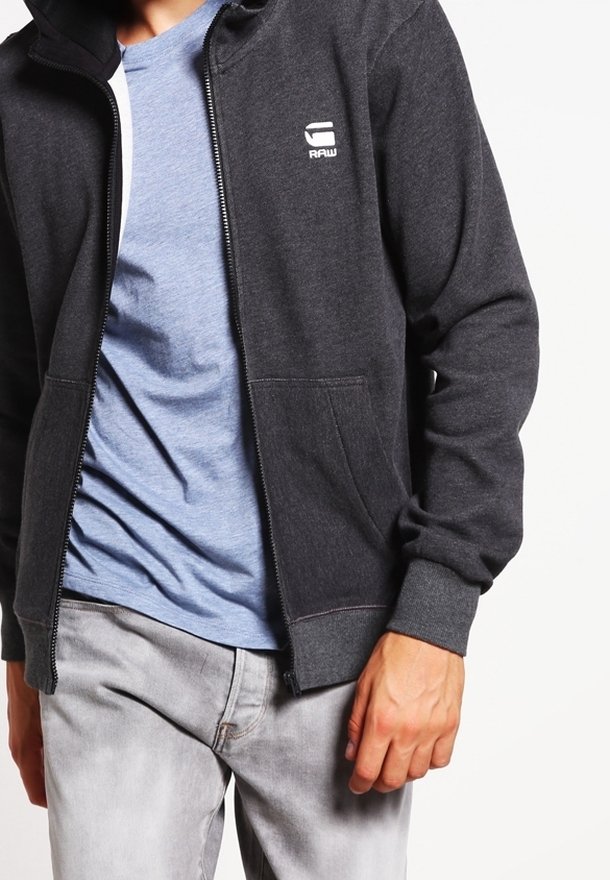 Homme portant un sweat à capuche zippé gris foncé avec un logo blanc sur la poitrine, par-dessus un t-shirt bleu clair et un jean gris clair.