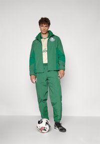 Grüne Sportjacke mit passender Hose, die ein Logo und Akzente aufweist. Das Outfit wird mit schwarzen Fußballschuhen und einem gemusterten Fußball kombiniert.