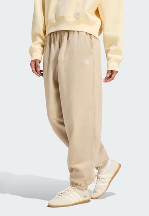 Beżowe dresy Adidas z elastycznymi ściągaczami przy kostkach i logo na udzie, noszone z pasującą bladożółtą bluzą i kremowymi butami Adidas.