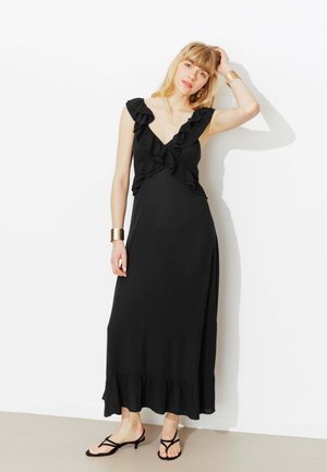 Femme en robe maxi noire sans manches avec des volants, portant des sandales noires à brides à talons bas et des bracelets en or, debout contre un mur blanc.