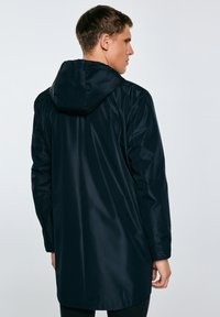 Vīrietis, kurš valkā garu melnu kapuci ar mēteļa kapuci, redzams no aizmugures pret vienkāršu gaišu fonu.