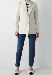 Blazer blanc texturé avec deux poches avant et deux boutons, assorti à un jean foncé et des chaussures à talons blocs noir et blanc.