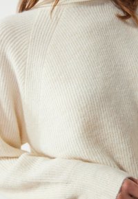 Pull en tricot côtelé crème avec col montant, présentant une texture douce et des poignets repliés. La vue rapprochée met en avant les détails et le motif du tissu.